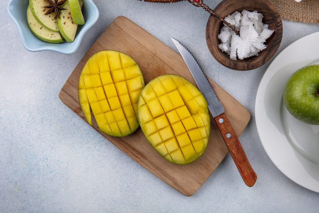 top-view-sliced-fresh-mango-wooden-kitchen-board-with-knife-chopped-apples-white-bowl-pulps-coconut-wooden-bowl-white-surface-scaled.jpg